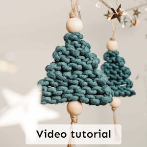 Op de afbeelding: Twee teal macramé kerstboomornamenten met houten kralen en jute touw om op te hangen. De ornamenten hebben de vorm van kerstbomen. De tekst "Video tutorial" wordt weergegeven op een witte rechthoek onderaan de afbeelding.