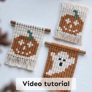 Könnte beinhalten: Drei Makramee-Wandbehänge mit Halloween-Motiven. Zwei zeigen orangefarbene Kürbisse mit grünen Stielen, und einer zeigt einen weißen Geist mit schwarzen Augen und Mund. Jedes Stück hat einen Holzstab und Fransen. Der Text "Video tutorial" ist sichtbar.