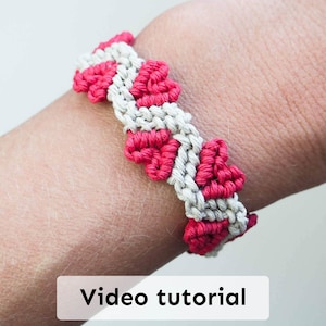 May include: A handmade macrame bracelet in a repeating pattern of red and off-white. The bracelet is worn on a wrist. The text "Video tutorial" is displayed in a white rectangle at the bottom of the image.