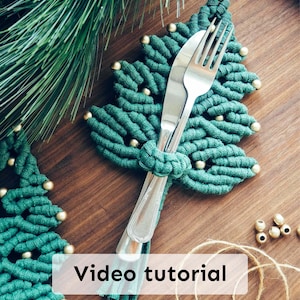 May include: A green macrame Christmas tree-shaped placemat with gold beads, holding a fork and knife. The text "Video tutorial" is displayed on the placemat. The placemat is on a wooden surface, with pine needles in the background.