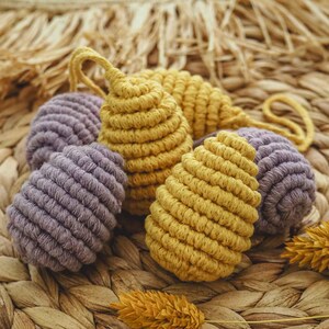 May include: A set of four crocheted Easter eggs in yellow and purple. The eggs are made with a macrame technique and are displayed on a woven wicker surface.