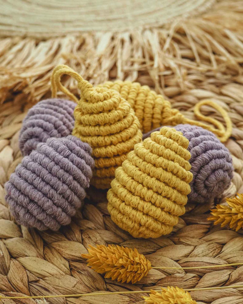 May include: A set of four crocheted Easter eggs in yellow and purple. The eggs are made with a macrame technique and are displayed on a woven wicker surface.