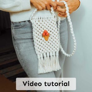 以下が含まれることがあります： 白いマクラメの携帯電話バッグ。織り込まれたアイスクリームコーンのデザイン。バッグには長いストラップと裾にフリンジが付いています。「Video tutorial」というテキストが画像の下部に表示されています。