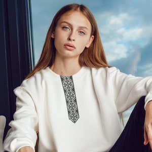 May include: A woman wearing a white sweatshirt with a black embroidered design, black pants, and black lace-up boots. She is sitting on a windowsill with a brick wall behind her.