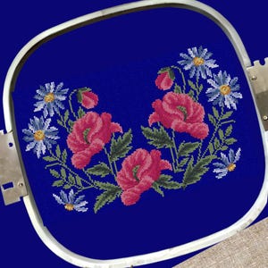 May include: Embroidered design featuring pink poppies and blue daisies on a dark blue fabric. The design is framed in a white embroidery hoop.