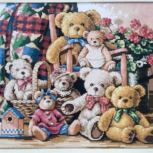 May include: A group of seven teddy bears in various poses, some sitting in a wicker basket, others sitting on a wooden chair. The bears are all different colours and have different expressions. There is a red, green and blue plaid blanket in the background.