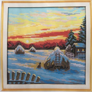 May include: A cross-stitch embroidery depicting a winter scene with snow-covered hay bales, a small house, and a pine tree in the foreground. The background features a vibrant sunset with orange, red, and yellow hues.
