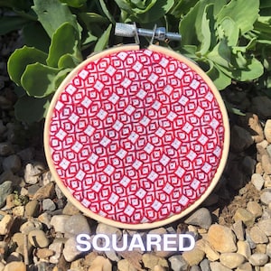 May include: A round embroidery hoop with a red and white geometric pattern. The word "SQUARED" is displayed at the bottom. The hoop is made of wood and has a metal tightening mechanism.