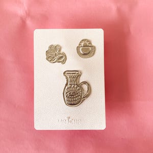 Puede incluir: Un conjunto de tres broches de metal plateado sobre una tarjeta blanca texturizada. Los broches incluyen una flor, un pequeño bolso y una jarra con un diseño de ojo. La tarjeta tiene el texto "MR. CRRP" impreso. El fondo es rosa.