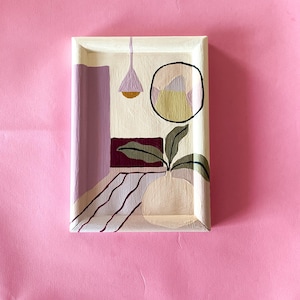 May include: Abstract painting on a rectangular wooden tray. The artwork features geometric shapes and a plant in a vase, with a cream, purple, and green colour palette. The background is a solid pink.