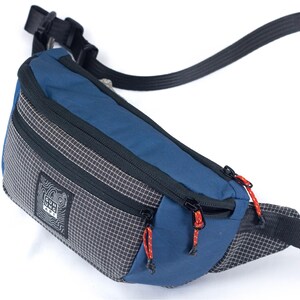 May include: A blue and gray fanny pack with black zippers and red pull cords. The bag has a checkered pattern on the sides and a black strap. The brand name "GUNG API" is printed on a white label.