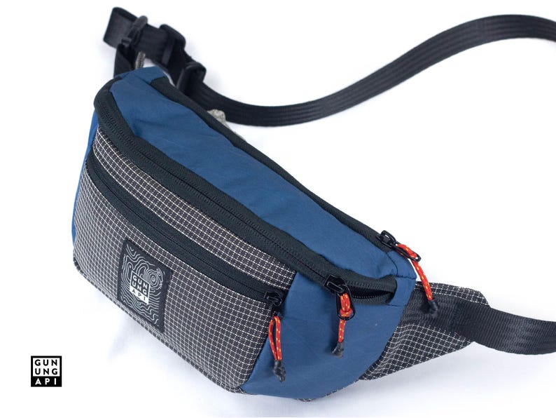 May include: A blue and gray fanny pack with black zippers and red pull cords. The bag has a checkered pattern on the sides and a black strap. The brand name "GUNG API" is printed on a white label.