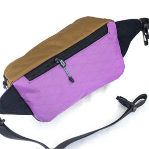 May include: A multi-colored fanny pack with a brown top, purple front, and black accents. The pack features a black zipper, a black adjustable strap, and a small logo on the bottom right corner. The fanny pack is designed for carrying essentials.