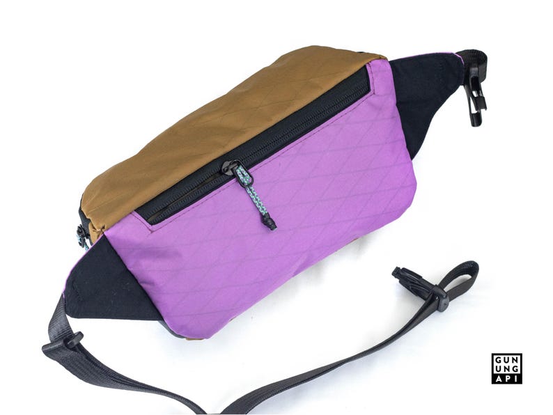 May include: A multi-colored fanny pack with a brown top, purple front, and black accents. The pack features a black zipper, a black adjustable strap, and a small logo on the bottom right corner. The fanny pack is designed for carrying essentials.