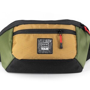 May include: A multi-colored fanny pack with a black base, olive green sides, and a tan front pocket. The front pocket has a black zipper with a red pull. A small square patch with a white graphic and text is centered on the front.