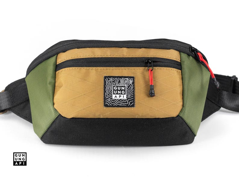 May include: A multi-colored fanny pack with a black base, olive green sides, and a tan front pocket. The front pocket has a black zipper with a red pull. A small square patch with a white graphic and text is centered on the front.