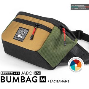May include: A multi-colored bum bag with the text "GUNUNG API JABOI 2.5L BUMBAG M / SAC BANANE". The bag features a black base, a tan front panel, and an olive green side panel. It has red zipper pulls and a black adjustable strap.