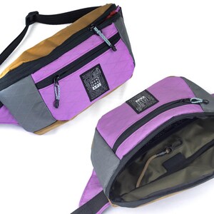 May include: Two fanny packs in a studio shot. One is open, revealing the interior. The bags are a mix of purple, gray, and tan with black zippers and straps. The brand logo is visible on the front of each bag.
