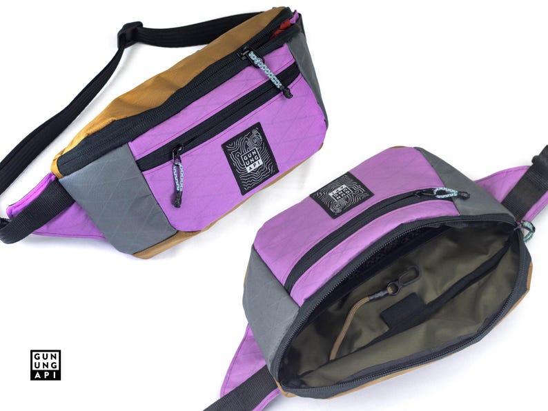 May include: Two fanny packs in a studio shot. One is open, revealing the interior. The bags are a mix of purple, gray, and tan with black zippers and straps. The brand logo is visible on the front of each bag.