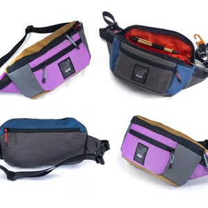 May include: A collection of colorful fanny packs in various angles. The bags feature a mix of colors including purple, blue, brown, and gray. Each bag has a black adjustable strap and a zippered compartment. One bag is open, revealing an orange interior and a pen.