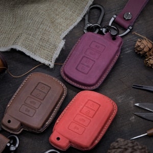 May include: Three leather key covers in different colors: brown, red, and purple. Each cover has four buttons and a stitched edge. The covers are on a wooden surface.