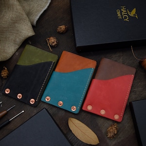 Könnte beinhalten: Vier Lederkartenhalter in verschiedenen Farben: Grün, Braun, Blau und Rot. Jeder Halter hat ein kontrastierendes Innenfutter und ist mit Kupfernieten befestigt. Die Halter sind auf einer Holzoberfläche mit einer kleinen Schachtel und einem Stück Stoff im Hintergrund angeordnet. Die Schachtel trägt den Schriftzug "HALCY CRAFT".