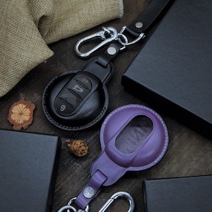 May include: Two leather keychains with key fobs. One keychain is black with a round key fob and the other is purple with a rectangular key fob. Both keychains have a silver metal hook.