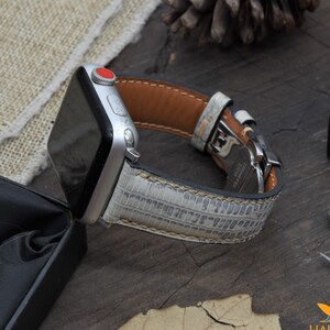 May include: A silver Apple Watch with a white leather band with a textured pattern. The band has a brown leather lining.