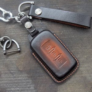 Op de afbeelding: Een bruine leren sleutelhanger met een gestikte rand en een zilveren sleutelring. De sleutelhanger heeft een ontwerp van een autosleutel met knoppen.