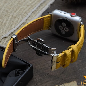 May include: A yellow leather watch band with a silver buckle and a silver Apple Watch.