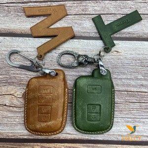 May include: Two leather key covers with a skull keychain. The key covers are brown and green with stitching around the edges. The key covers have three buttons on the front. The keychain has a skull charm and a silver ring.