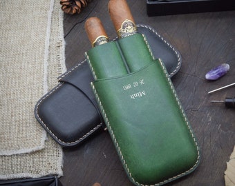 Puede incluir: Un estuche de cuero verde para cigarros con dos cigarros en su interior. El estuche tiene una inscripción personalizada que dice "Minh 26.02.1993".