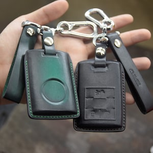 Może przedstawiać: Dwa skórzane breloki z karabińczykiem ze srebrnego metalu. Jeden brelok jest czarny ze szwem w kolorze zielonym i zielonym kółkiem w środku. Drugi brelok jest czarny ze szwem w kolorze brązowym i srebrnym tłoczonym wzorem. Brelok z zielonym kółkiem ma wytłoczone na pasku słowo "CROSSOVER".