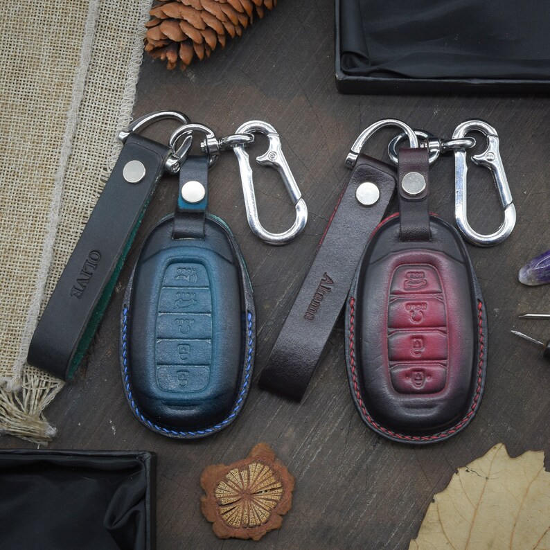 May include: Two leather keychains with metal key rings and car key covers. One keychain is brown with the word "Altitude" embossed on it and the key cover is red. The other keychain is black with the word "Olive" embossed on it and the key cover is blue.