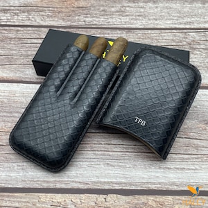 Könnte beinhalten: Schwarzes Leder-Zigarrenetui und -halter mit strukturiertem Schlangenhautmuster. Der Halter enthält drei Zigarren. Das Etui ist mit den Initialen "TPB" geprägt. Eine schwarze Box befindet sich im Hintergrund.