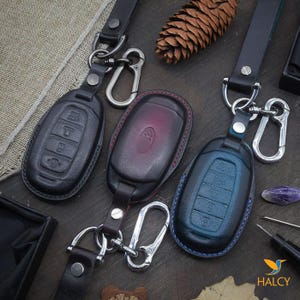 May include: Three leather keychains with different colored key covers. The key covers are black, burgundy, and blue. The keychains have silver metal rings and hooks.