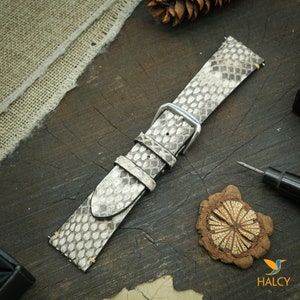 Pode incluir: Uma pulseira de rel&oacute;gio de pele de cobra branca com uma fivela prateada. A pulseira est&aacute; sobre uma superf&iacute;cie de madeira.