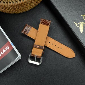 May include: A brown leather watch strap with a textured pattern and a light brown underside. The strap is folded in half and the buckle is visible.