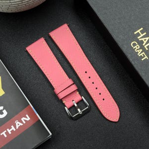 May include: Two pink leather watch straps with yellow stitching. The straps are laid out on a black surface.