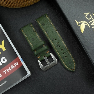 May include: Two green leather watch straps with orange stitching. The straps have an embossed pattern and are shown on a black surface.