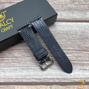 Peut inclure: Deux bracelets de montre bleu marine avec un motif texturé en forme de reptile. Les bracelets ont des coutures beige clair et des boucles argentées. Les bracelets sont présentés sur une surface en bois à côté d'une boîte noire avec l'inscription dorée "HALCY CRAFT".