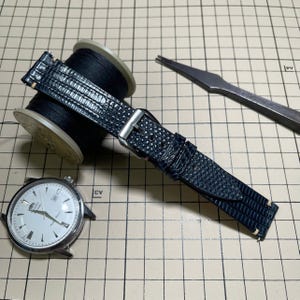 Könnte beinhalten: Ein dunkelblaues Uhrenarmband mit texturiertem Eidechsenhautmuster und einer silberfarbenen Metallschnalle. Das Armband liegt neben einer runden silbernen Armbanduhr mit weißem Zifferblatt, einer Spule mit schwarzem Garn und einem Metallwerkzeug.
