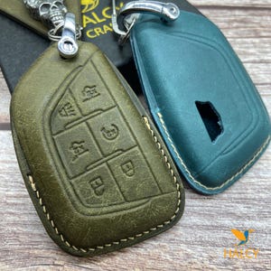 May include: Two leather key covers, one in dark green and one in teal blue. Both covers have a stitched edge and are designed to fit a car key fob. The green cover has a textured surface and embossed buttons.