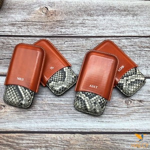 Pode incluir: Quatro estojos de charuto de couro marrom com um padrão de pele de cobra no interior. Os estojos são personalizados com os nomes "NRJJ", "TL", "AJAY" e "LCFB".