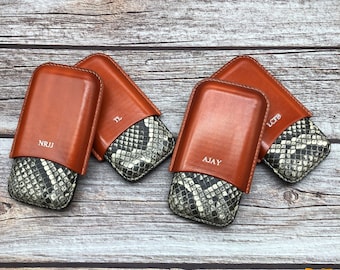 Puede incluir: Cuatro estuches para cigarros de cuero marrón con un patrón de piel de serpiente en el interior. Los estuches están personalizados con los nombres "NRJJ", "TL", "AJAY" y "LCFB".