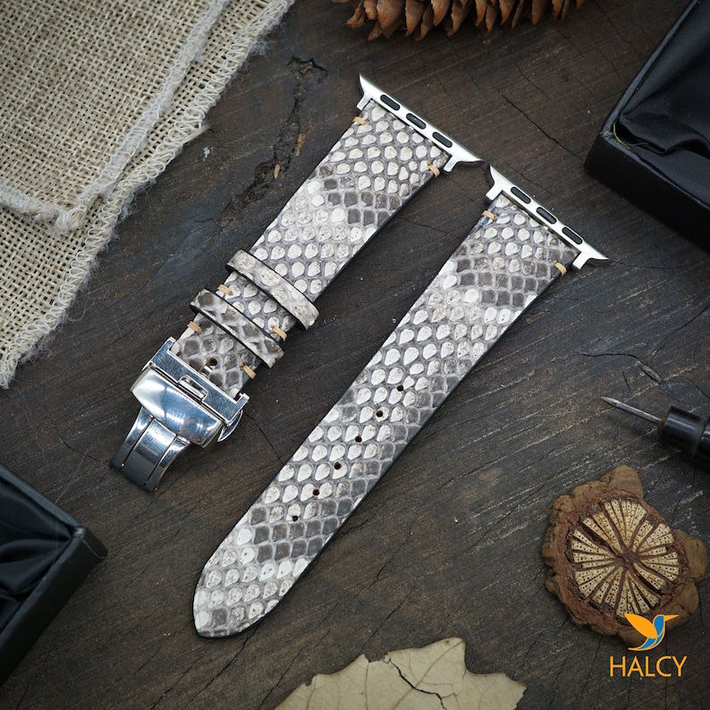 May include: A gray and white snakeskin Apple Watch band with a silver buckle. The band is made of genuine leather and has a classic design.