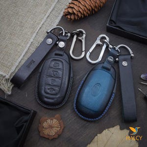 May include: Two leather keychains with key fob covers. One keychain is black with a blue key fob cover and the other is black with a black key fob cover. Both keychains have a silver metal loop and a black leather strap.