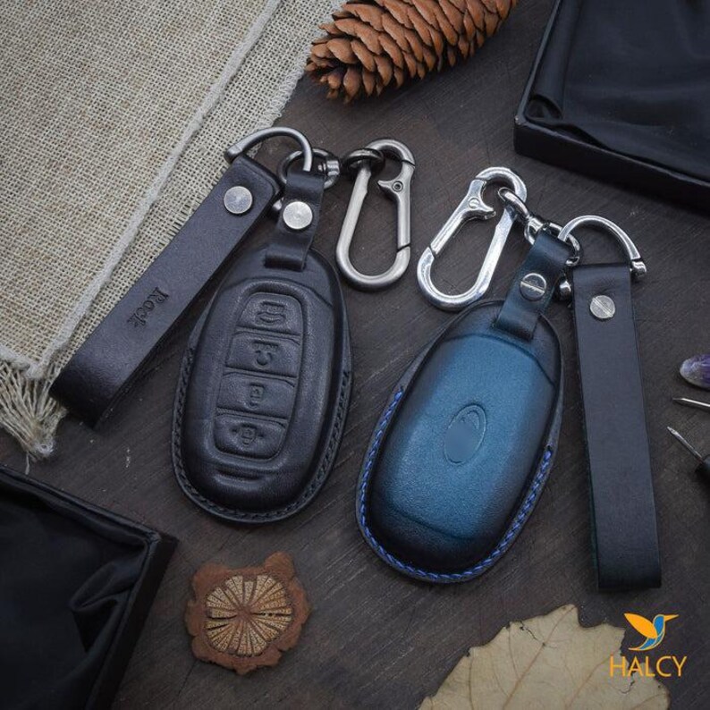 May include: Two leather keychains with key fob covers. One keychain is black with a blue key fob cover and the other is black with a black key fob cover. Both keychains have a silver metal loop and a black leather strap.