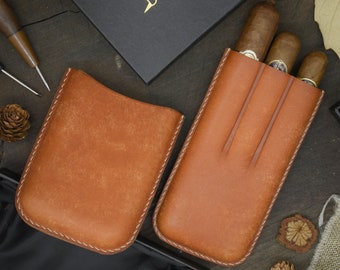 May include: Two brown leather cigar cases with stitched edges. One case is empty and the other case holds three cigars.