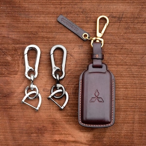 Może przedstawiać: Dwa srebrne breloki do kluczy z kółkiem D i haczykiem oraz brązowy skórzany brelok do kluczy ze złotym zapięciem i logo Mitsubishi. Brelok do kluczy ma napis "USS410" z tyłu.